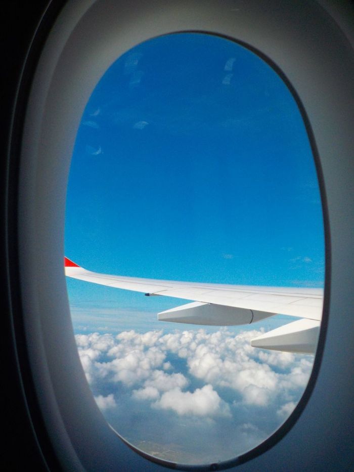 social-feed-img-02 View of an airplane wing and fluffy clouds through a plane window, with a bright blue sky.